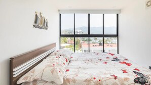 A bedroom with a large window, a bed with floral bedding, a wooden headboard, and a wall-mounted light fixture. - Departamento Astoria (Acapulco de Juarez)