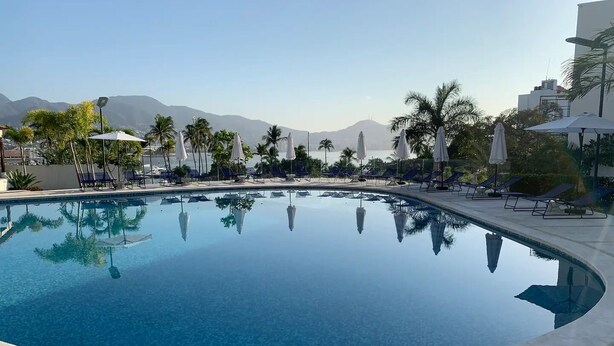 A clear swimming pool with lounge chairs and umbrellas, surrounded by palm trees and mountains in the background. - Departamento Astoria (Acapulco de Juarez)