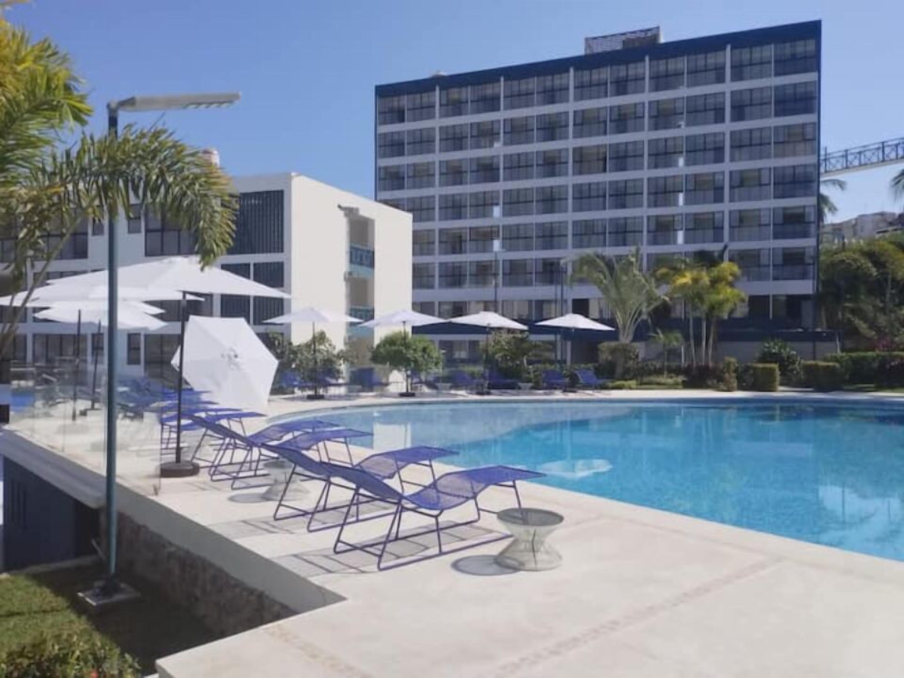 Una piscina amb gandules i ombrel·les, envoltada de palmeres i edificis moderns.