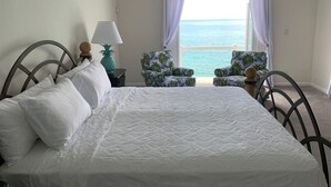 A bedroom with a bed, two chairs, a nightstand, a lamp, and a view of the ocean.