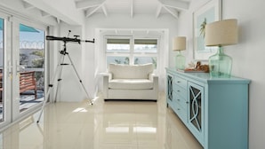 A room with a white sofa, a teal cabinet, a telescope, and a window with a view of the outside.