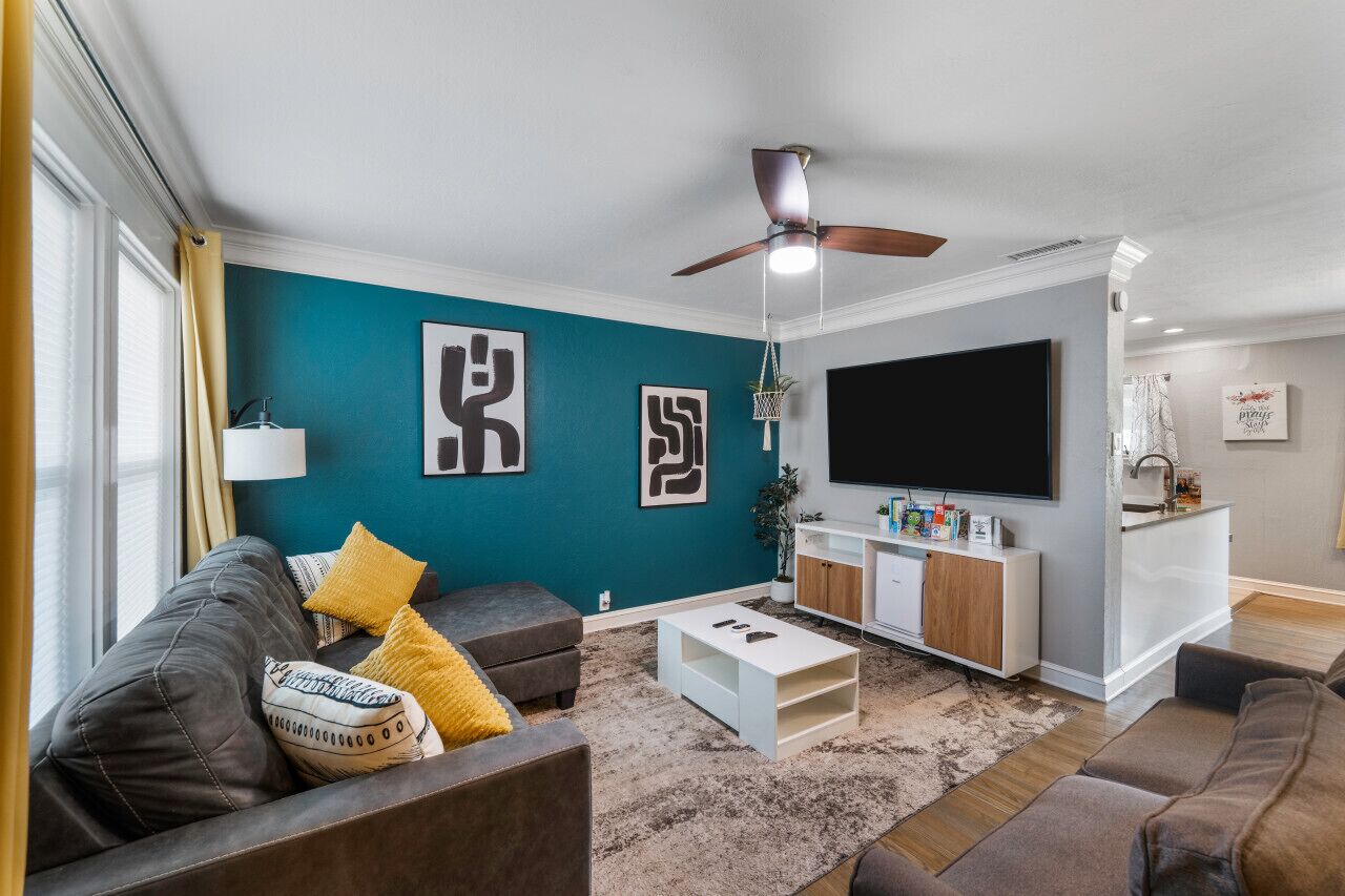 A modern living room with teal walls, a ceiling fan, a flat-screen TV, a coffee table, and a sofa with cushions.