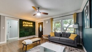A living room with a grey sofa, a white coffee table, a ceiling fan, and a wall with a neon sign.
