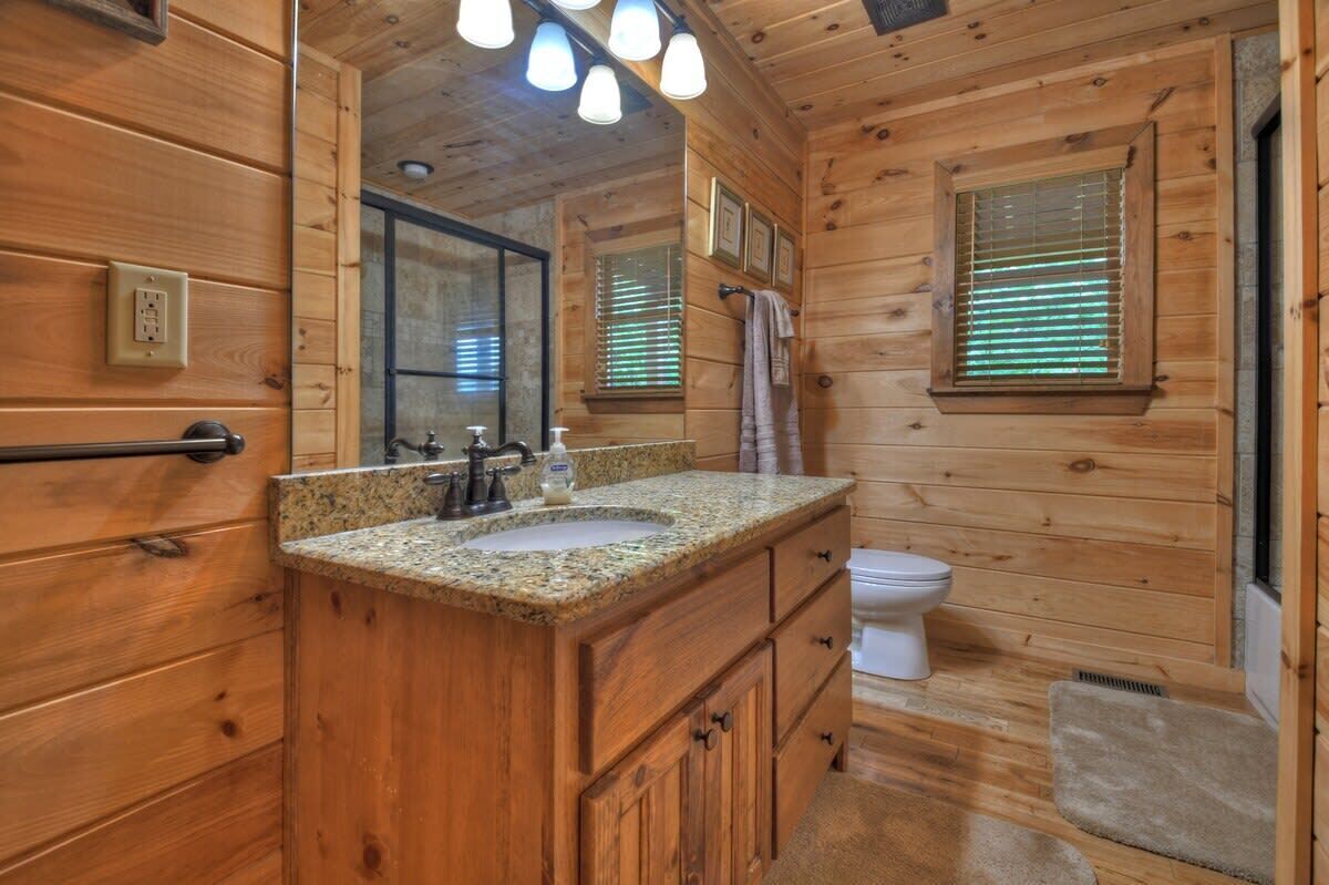 A wooden cabin bathroom with a granite countertop sink, a white toilet, and a window with blinds.