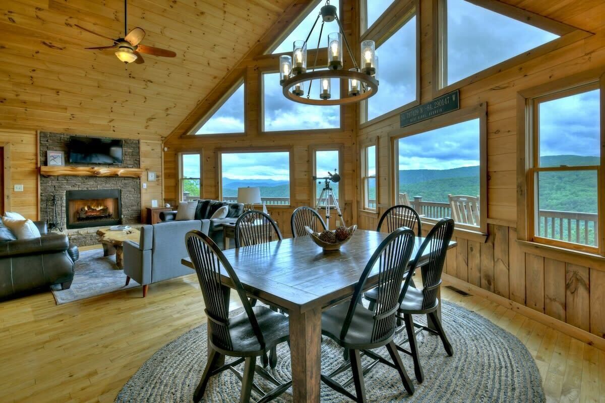A wooden cabin interior with a fireplace, dining table, chairs, and large windows offering a view of the mountains.