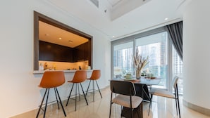 A modern dining area with a kitchen in the background, a large window with a view of tall buildings, and a table set with chairs. - EDEN'S Homes & Villas - Opera Grand (Dubai)