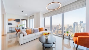 A modern living room with a white sofa, orange armchair, and a round coffee table with fruit. A city view is visible through large windows. - EDEN'S Homes & Villas - Opera Grand (Dubai)