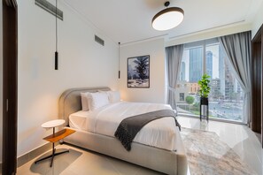 A modern hotel room with a large bed, a small side table, a potted plant, and a city view through the window.