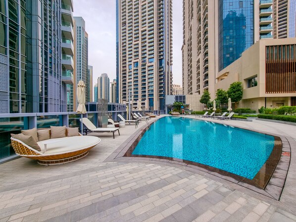 A rooftop swimming pool with lounge chairs and a wicker sofa, surrounded by high-rise buildings.