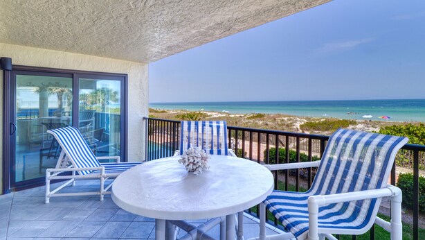 A balcony with two striped lounge chairs, a round table with a vase, and a view of the beach and ocean.