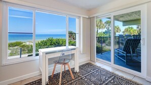 A room with a desk and chair, large windows with a view of the beach, and a balcony with outdoor furniture.