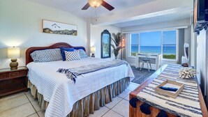 A hotel room with a bed, a desk, a chair, a ceiling fan, a large window with a view of the ocean, and a painting on the wall.
