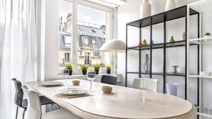 A dining area with a table set for two, a window with a view of buildings, and a black bookshelf with decorative items. - Muette - Lamartine 1bdr by B'Your Home (Paris)