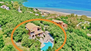 An aerial view of a coastal property with a swimming pool, surrounded by lush greenery and a beach.
