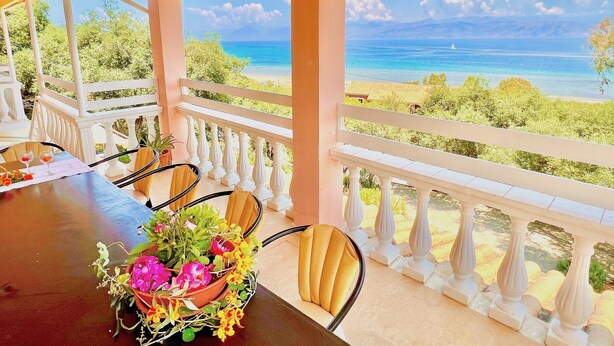 A covered outdoor seating area with a table set for two, a flower centerpiece, and a view of the ocean and beach.