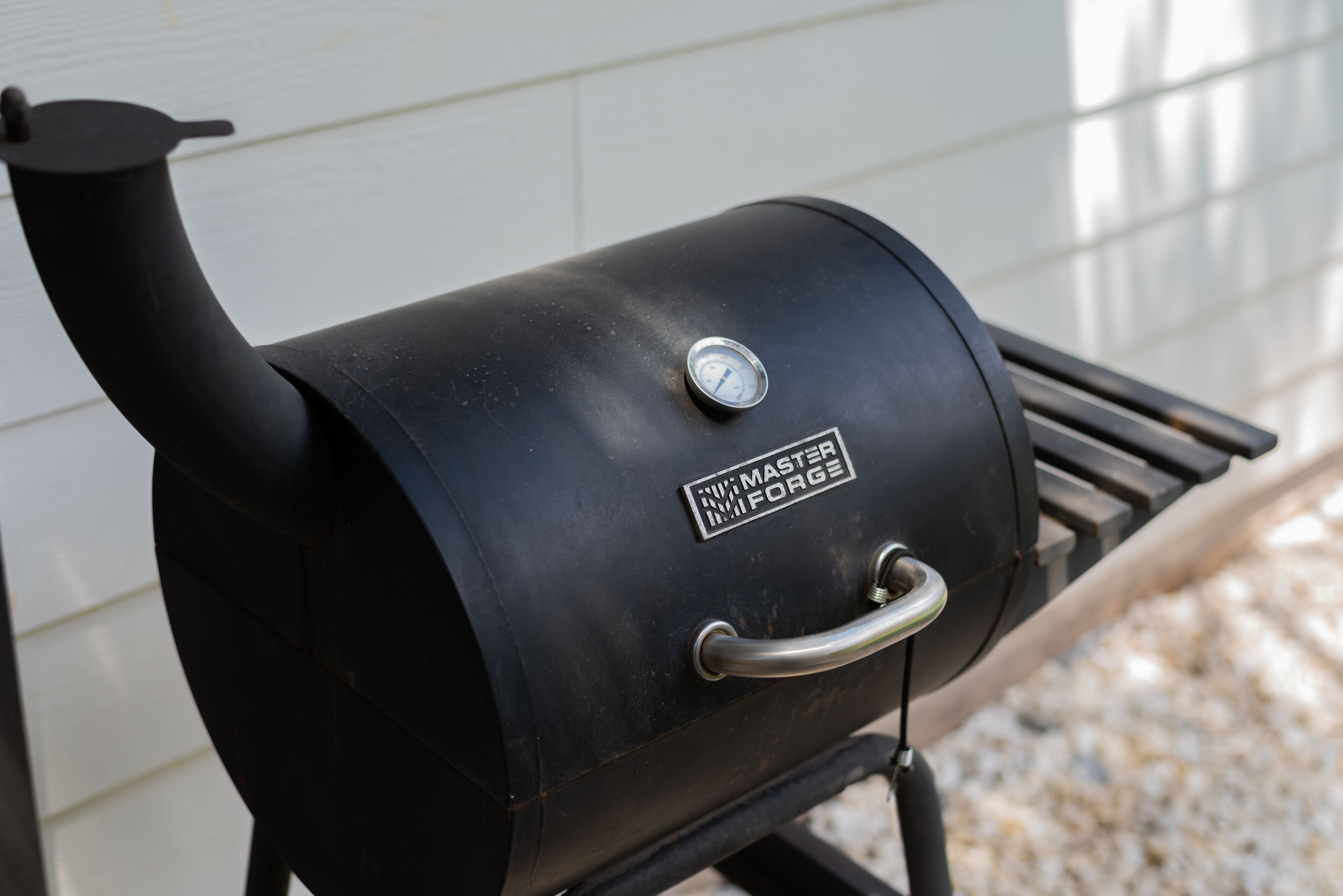 A black outdoor grill with a pressure gauge and a handle, placed on a grill rack against a white wall.