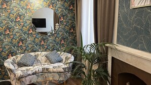 Una habitación con papel tapiz estampado, una mesa redonda con un espejo, un sofá, una planta en maceta y una chimenea.
