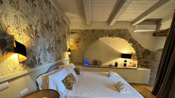 Un dormitorio con pared de piedra, una cama con ropa de cama blanca, una mesita de noche con teléfono y una lámpara de pared.