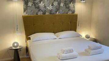 A neatly made bed with white linens and rolled towels, a mustard-colored headboard, and a floral-patterned wall above.