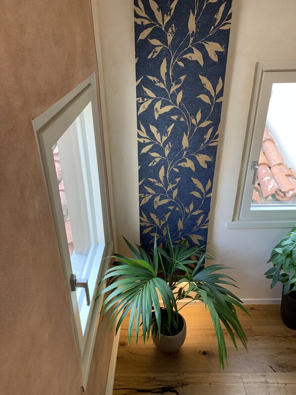 Una habitación con piso de madera, una planta en maceta, pared con diseño azul y una ventana con vista a un tejado de tejas rojas.