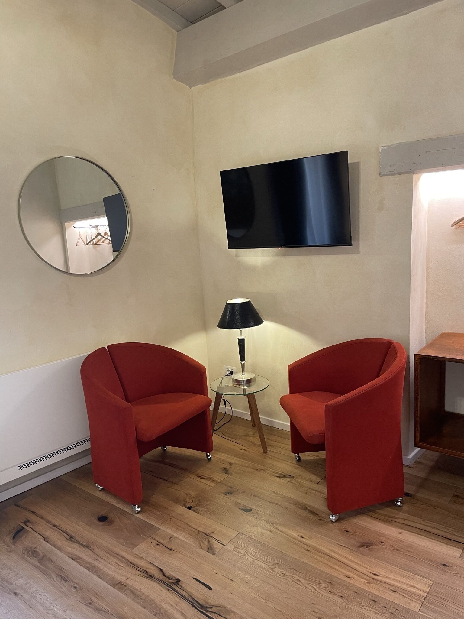 Una habitación con dos sillones rojos, un espejo redondo, una mesita con una lámpara y un televisor de pantalla plana montado en la pared.