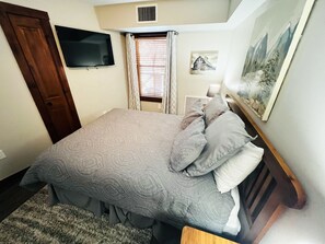 A bedroom with a bed, a television, a painting, and a window with curtains.