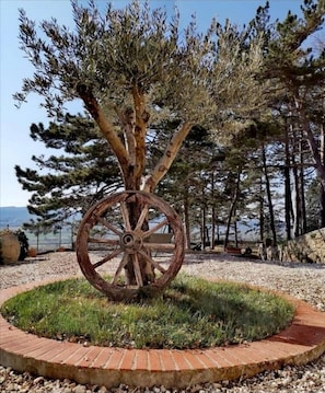 A rustic wooden wheel sculpture surrounded by a circular brick pathway, set against a backdrop of pine trees and a clear sky. - Dream House (Arezzo)