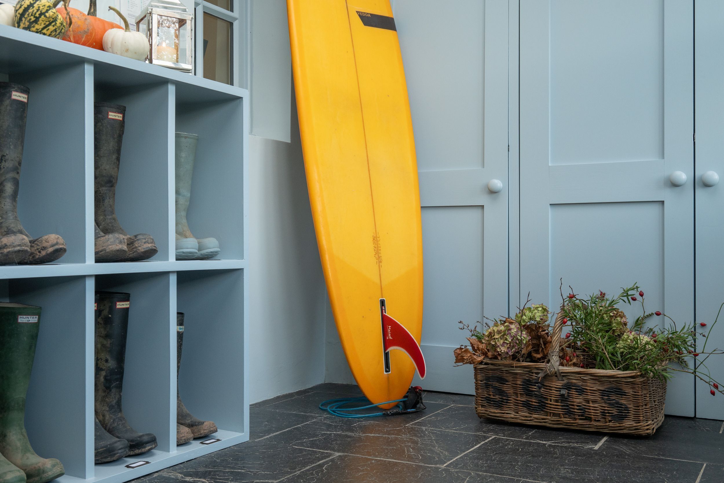 Una tabla de surf apoyada en una pared, un estante con botas y una cesta con plantas secas.