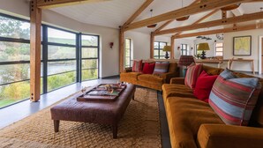 Un vaste salon doté de grandes fenêtres, d’un plafond en bois et d’un coin salon confortable avec un canapé et un pouf marron.
