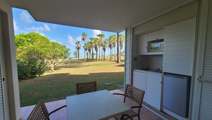 Um pátio com mesa e cadeiras, uma pequena cozinha com geladeira e vista para uma área gramada com palmeiras.