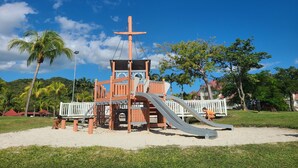 A playground with a slide and climbing structures, surrounded by palm trees and a clear blue sky. - Studio Résidence Pierre et Vacances 4 Personnes (Sainte-Luce)