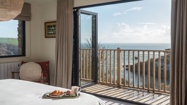 A bedroom with a large window overlooking a coastal view, a bed with a breakfast tray, and a painting on the wall. - Murrawingi -  a beautiful modern coastal home (Salcombe)