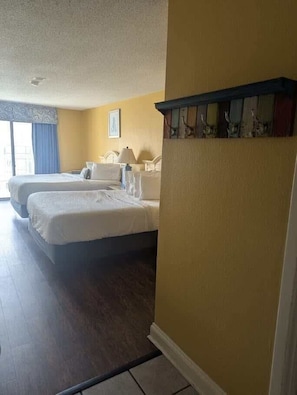 Una habitación de hotel con dos camas, una ventana con cortinas, un estante en la pared con objetos decorativos y una puerta.