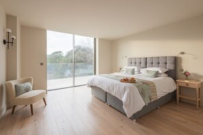 Ein geräumiges Schlafzimmer mit einem großen Bett, einem Sessel, einem Nachttisch und Blick ins Freie durch eine Schiebetür.