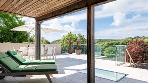 Eine Dachterrasse mit grünen Liegestühlen, einem Glas-Swimmingpool und Blick auf die umliegende Landschaft.