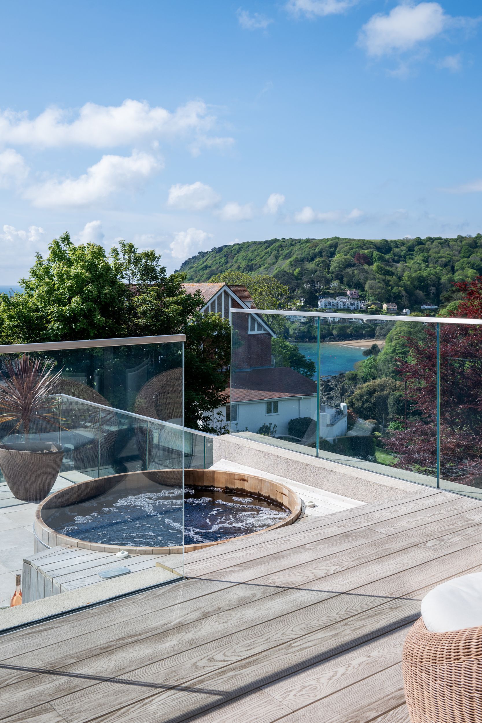 Una terraza de madera con jacuzzi, barandilla de vidrio, mobiliario de mimbre y una vista panorámica de una zona costera con colinas y edificios.