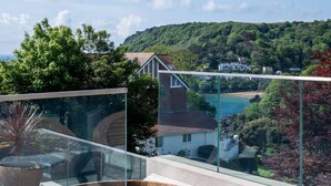 Eine Holzterrasse mit Whirlpool, Glasgeländer, Korbmöbeln und einem herrlichen Ausblick auf eine Küstenlandschaft mit Hügeln und Gebäuden.