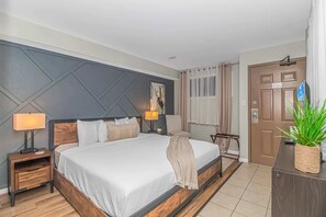 A hotel room with a large bed, bedside lamps, a wooden nightstand, a small table, a potted plant, and a door with a number.