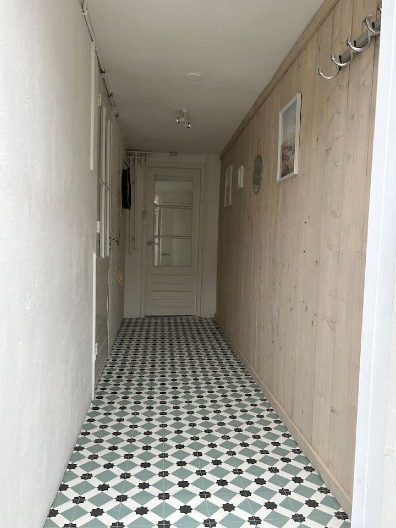 A narrow hallway with patterned floor tiles, a wooden wall, and a white door at the end.