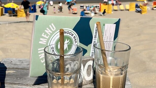 Ein Holztisch mit zwei Gläsern eines braunen Getränks, einer Speisekarte und einem Strand im Hintergrund.