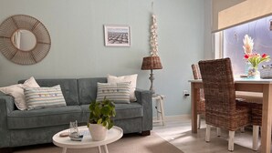 Ein Wohnzimmer mit grauem Sofa, rundem Couchtisch mit Pflanze, Korbsessel und Esstisch mit Stühlen.