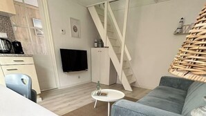 Ein modernes Wohnzimmer mit einer Couch, einem Couchtisch und einem Esstisch. Eine Treppe mit weißer Geländer, ein an der Wand montierter Fernseher und ein Korbstuhl sind ebenfalls vorhanden.