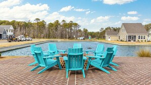 Un patio con cuatro sillas Adirondack turquesa alrededor de un hogar a leña de ladrillo, con vista a una piscina y un lago, con casas al fondo.