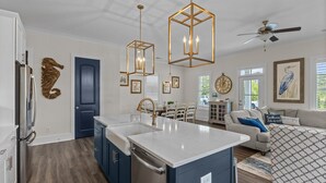 Una cocina moderna con una isla central, un comedor con mesa y sillas, una sala de estar con sofá y cojines, y un ventilador de techo.