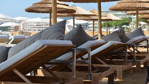 A row of wooden sun loungers with grey cushions, arranged on a sandy beach with thatched umbrellas in the background. - Luxe Appartement met 2 Slaapkamers, Groot Terras Direct aan Zwembad La Mourre 16 (Le Plan-de-la-Tour)