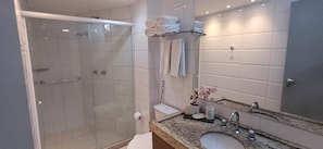 A modern bathroom with a glass-enclosed shower, a toilet, a sink with a marble countertop, and a towel rack with neatly folded towels. - Imperial Suítes (João Pessoa)