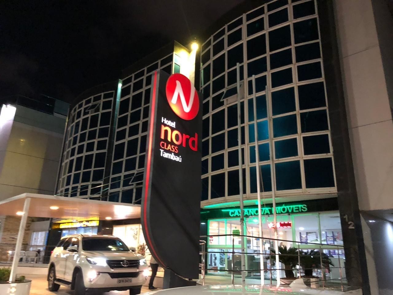 Entrada de hotel com uma placa chamativa para 'Nord Class Tambatu', fachada de vidro com a inscrição 'CASA MÓVETS' e um veículo branco estacionado na frente.