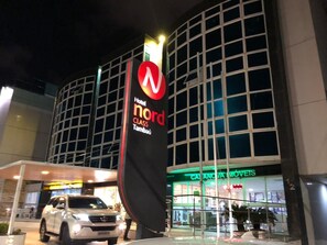 A hotel entrance with a prominent sign for 'Nord Class Tambatu,' a glass storefront with 'CASA MÓVETS' signage, and a white vehicle parked in front. - Imperial Suítes (João Pessoa)
