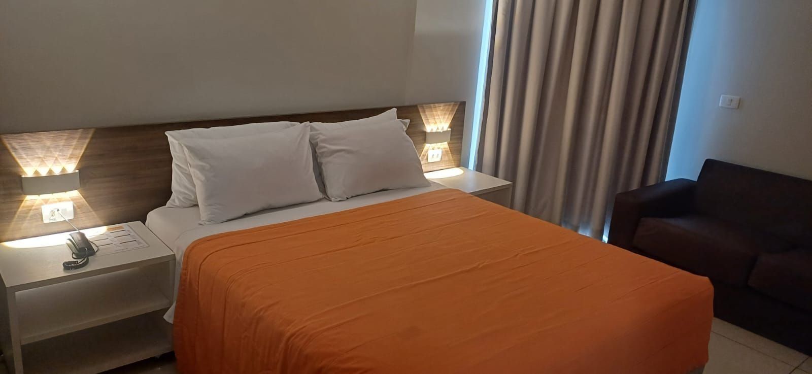 A hotel room with a bed, two bedside lamps, a nightstand, a remote control, and a brown leather chair.