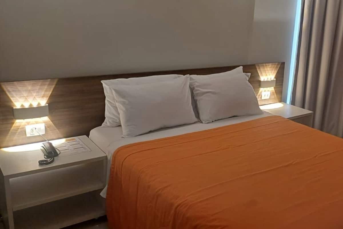 A hotel room with a bed, two bedside lamps, a nightstand, a remote control, and a brown leather chair.